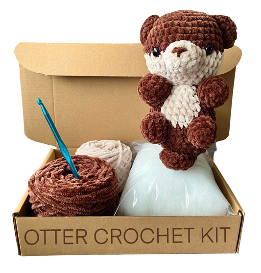 Completed crocheted otter on top of an open cardboard box including two colours of chenille yarn, toy stuffing and a crochet hook