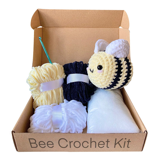 Crocheted bee on top of an open cardboard box next to three colours of chenille yarn, a crochet hook and polyester stuffing