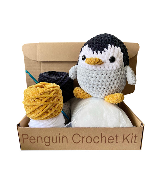 Finished handmade penguin on top of an open cardboard box next to chenille yarn, hook and stuffing