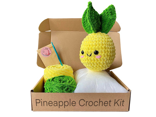 Crocheted pineapple on top of an open cardboard box besides two colours of chenille yarn (yellow and green), toy stuffing and a crochet hook