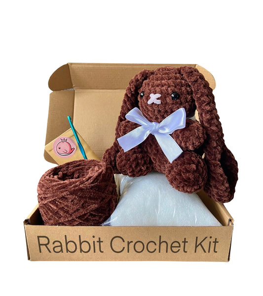 Crocheted rabbit on top of an open cardboard box including toy stuffing, chunky brown chenille yarn and a crochet hook