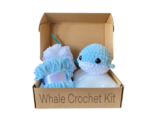 Crocheted whale on top of an open cardboard box including blue and white chenille yarn, toy stuffing and a crochet hook