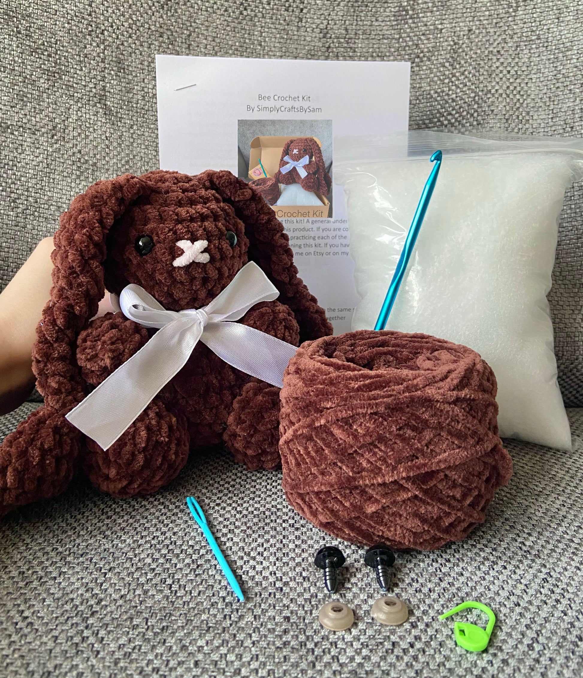 Finished crocheted bunny rabbit next to chunky chenille yarn, a stitch marker, safety eyes with backs, crochet hook, written crochet pattern and toy stuffing